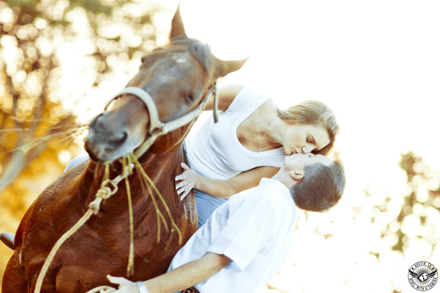 blond girl in white tank top and blue jeans riding on horde leans over and kisses guy in white short sleeve button up shirt with blurred trees in background in this engagement photo in south texas